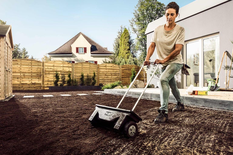 Eine Frau verwendet einen Streuwagen, um Rasensamen im Garten zu verteilen.