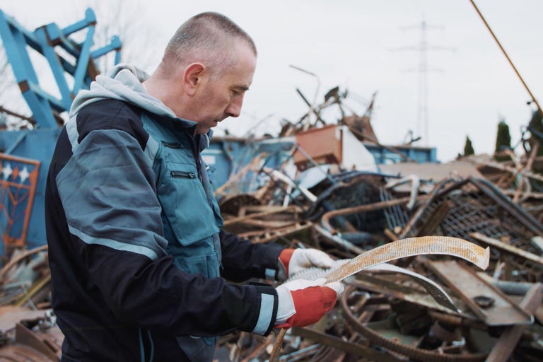 Mann in Arbeitskleidung begutachtet Metallschrott vor Schrottplatz