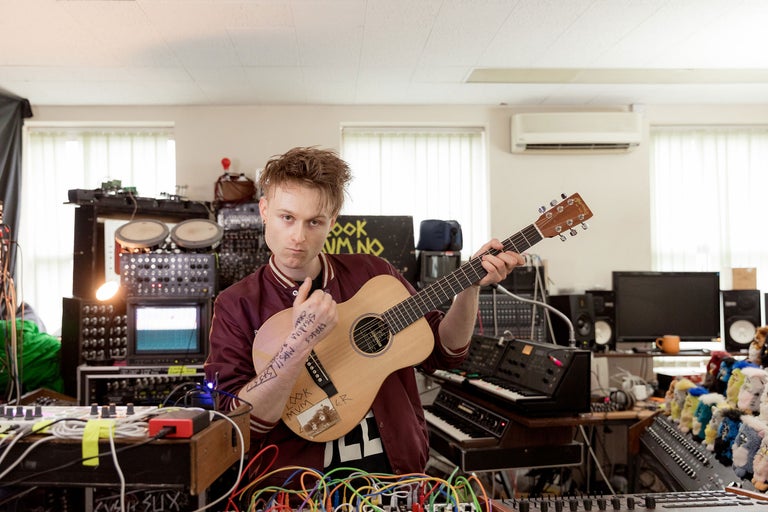 Ein Mann hält eine Akustikgitarre in einem Musikstudio mit Synthesizern und Mischpulten.