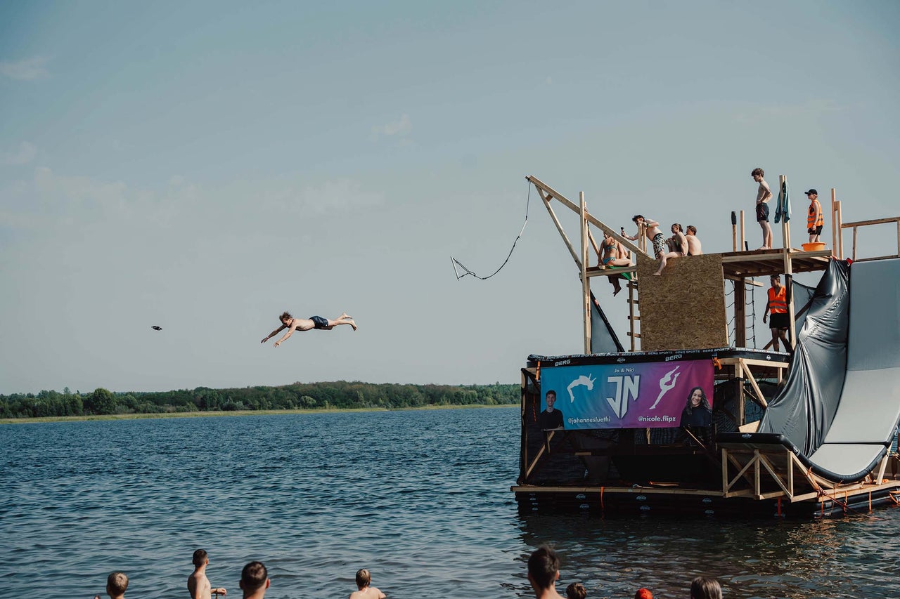 Wasserszene mit einer Gruppe von Personen auf einer schwimmenden Plattform und einer Person, die ins Wasser springt.