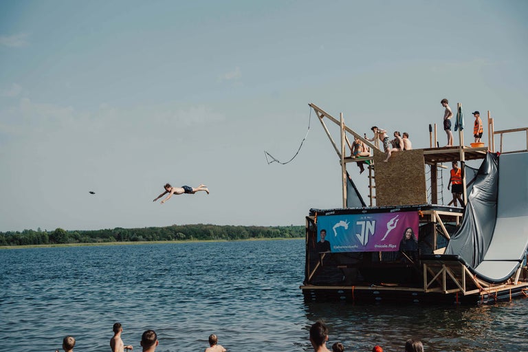 Wasserszene mit einer Gruppe von Personen auf einer schwimmenden Plattform und einer Person, die ins Wasser springt.