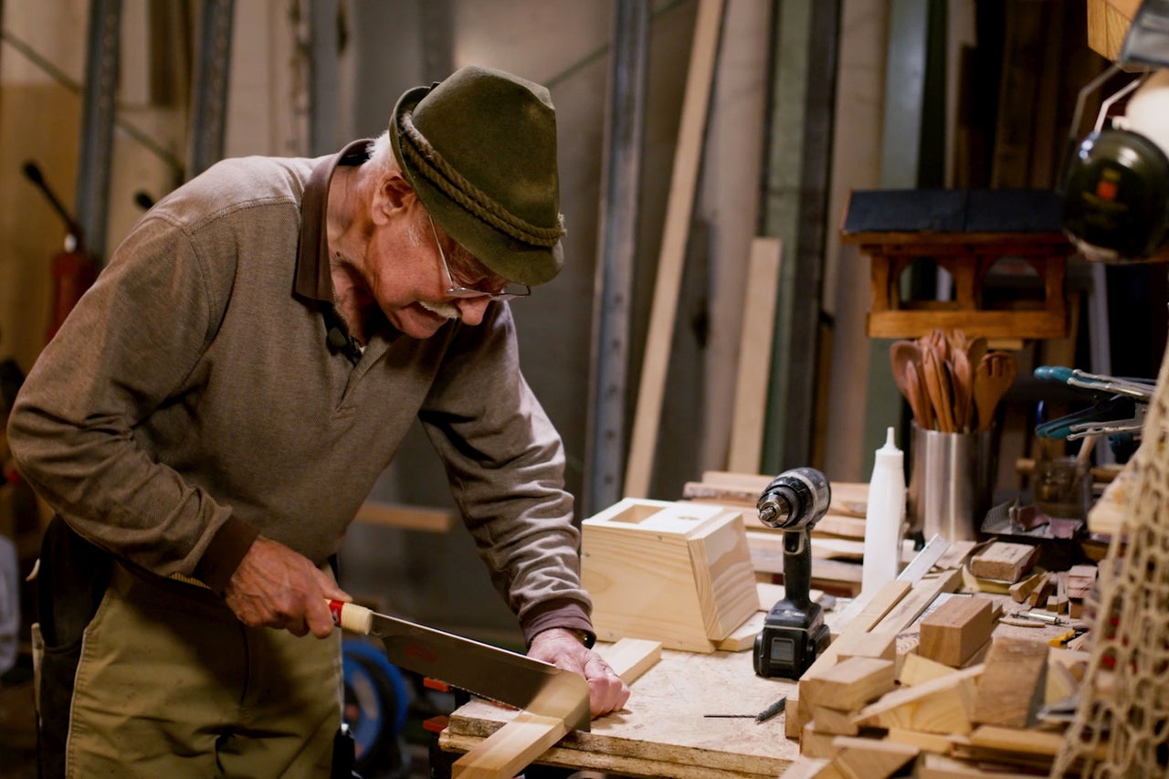 Ein Mann sägt Holz in einer Werkstatt mit einer Handsäge, umgeben von Werkzeugen und Materialien.