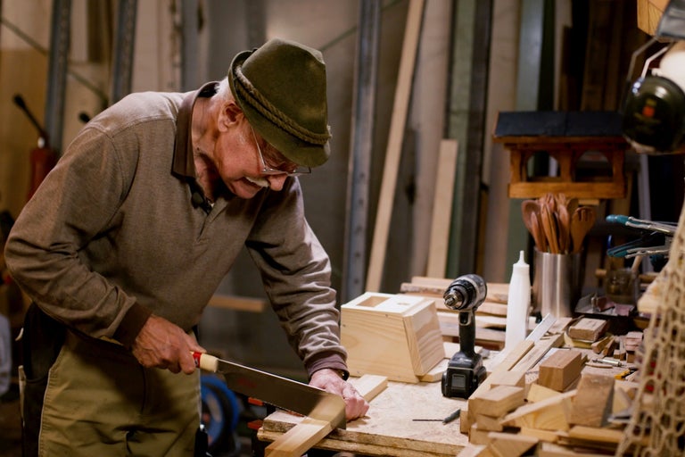 Ein Mann sägt Holz in einer Werkstatt mit einer Handsäge, umgeben von Werkzeugen und Materialien.