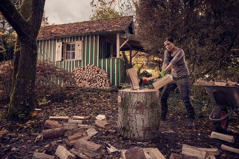 Eine Frau spaltet Holz mit einer Axt vor einem Holzschuppen in einem Garten.