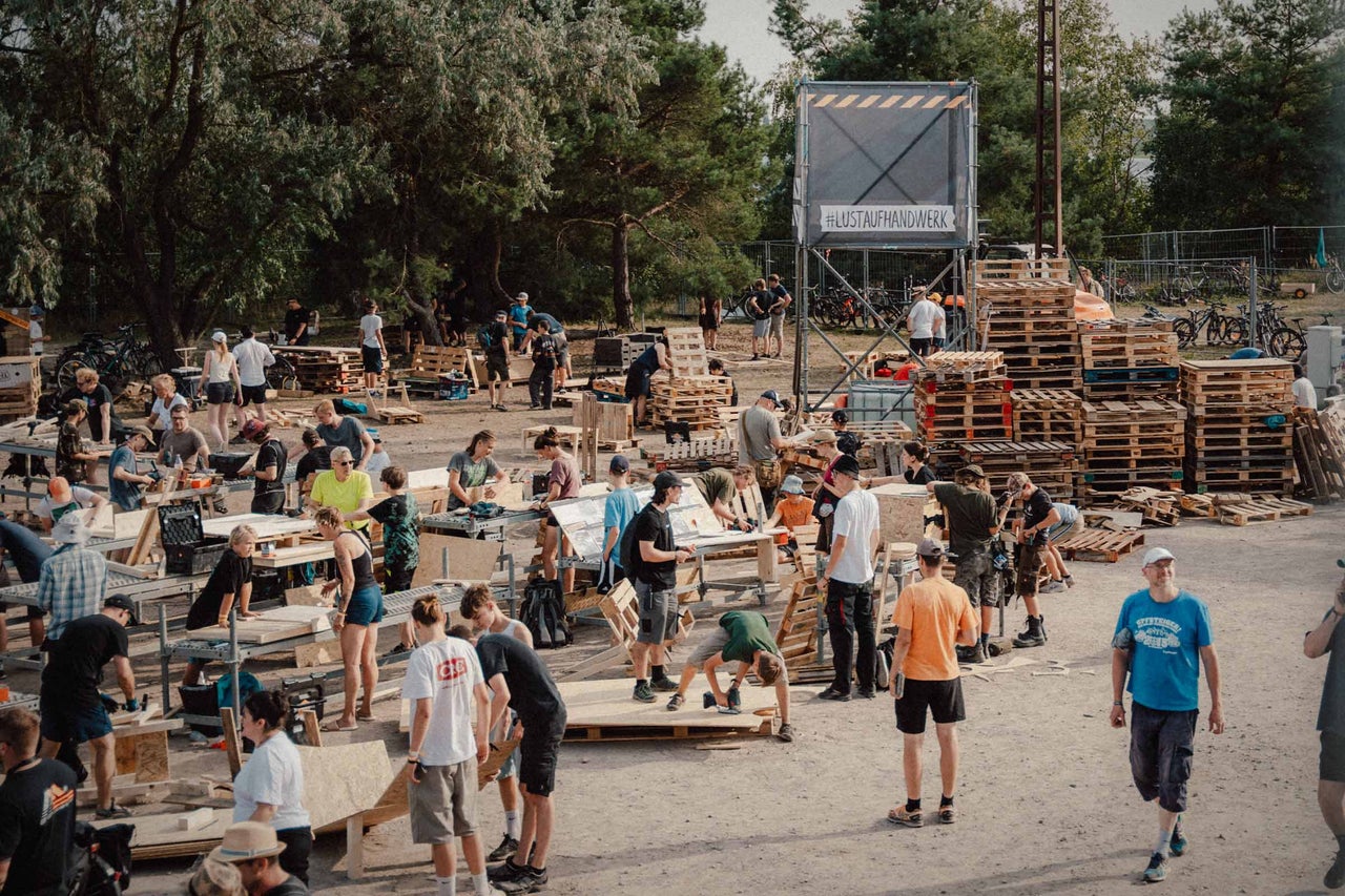 Eine Gruppe von Menschen arbeitet im Freien mit Holzpaletten und Werkzeugen, umgeben von Bäumen und einem Gerüst mit der Aufschrift '#LUSTAUFHANDWERK'.