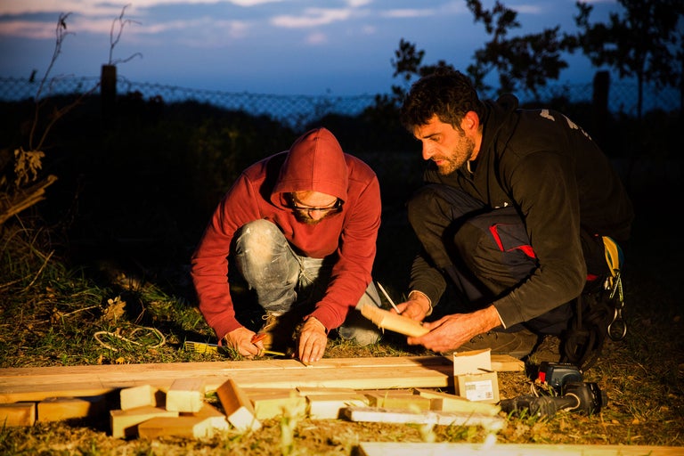Zwei Personen arbeiten mit Holz im Freien, wobei Werkzeuge wie Maßband und Stift verwendet werden.