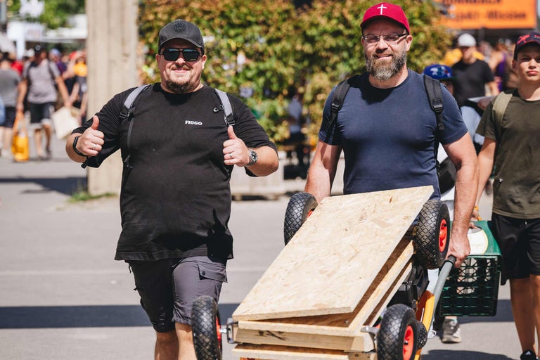 Zwei Männer mit Rucksäcken ziehen einen Bollerwagen mit OSB Platten bei einem Event unter freiem Himmel.