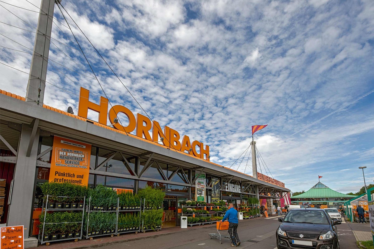  Außenansicht eines Hornbach Baumarktes mit Pflanzen vor dem Eingang und einem Kunden mit Einkaufswagen. 