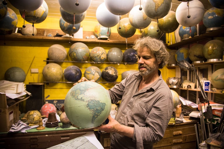 Ein Mann hält einen Globus in einem Raum mit zahlreichen Globen an der Decke und in Regalen.