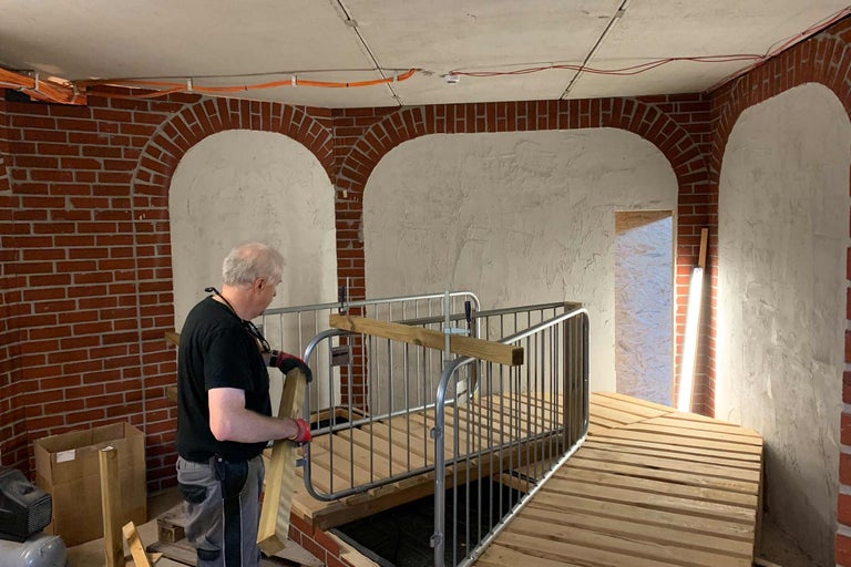 Mann montiert eine Rampe mit Geländer in einem Raum mit Mauerwerk und Putzwänden.
