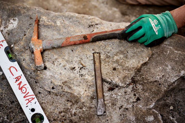 Werkzeuge auf Steinplatte: Spitzhacke, Meißel, Wasserwaage und Arbeitshandschuh.