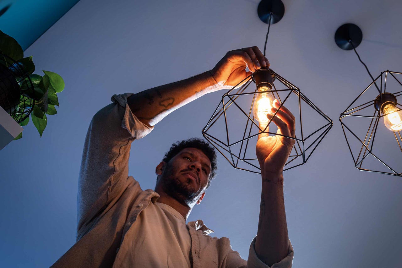 Ein Mann dreht ein leuchtendes LED-Leuchtmittel in eine schwarze, geometrische Hängeleuchte im modernen Design ein.