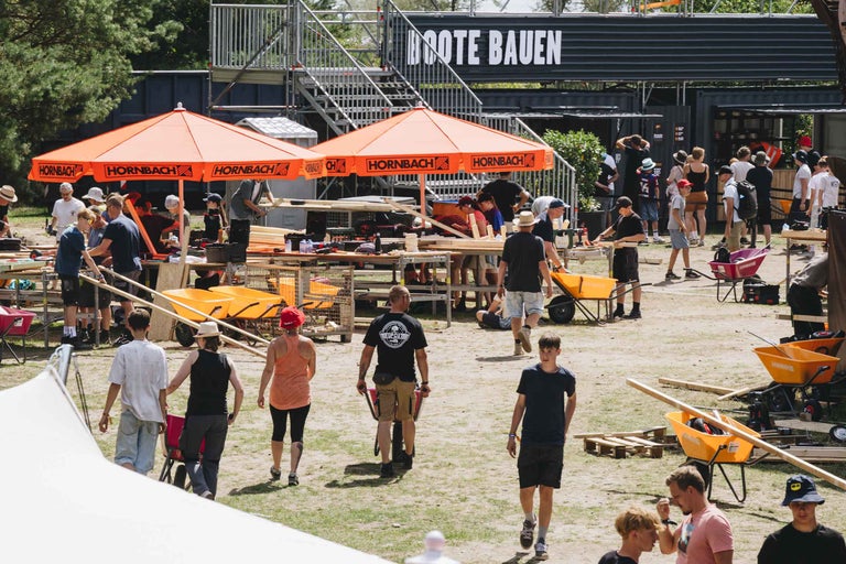 Menschen bauen an Werkbänken unter Hornbach Sonnenschirmen im Freien bei einem Event mit Material und Schubkarren.