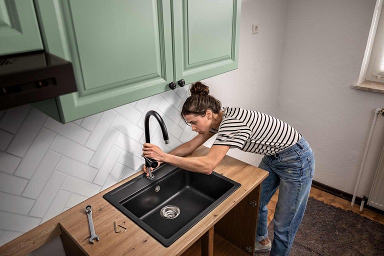 Eine Frau montiert eine schwarze Küchenarmatur an einem dunklen Einbauspüle in einer Küche mit hellen Fronten und Holz Arbeitsplatte.