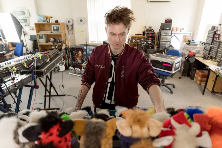 Szene eines Musikstudios mit elektronischen Musikinstrumenten und einer Person, die vor einer Sammlung von Fellpuppen steht.