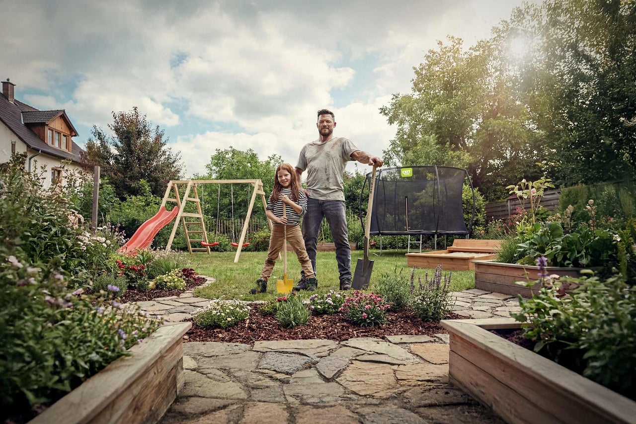 Gartenansicht mit Trampolin, Schaukel, Rutsche, Sandkasten, Vater und Tochter mit Gartengeräten