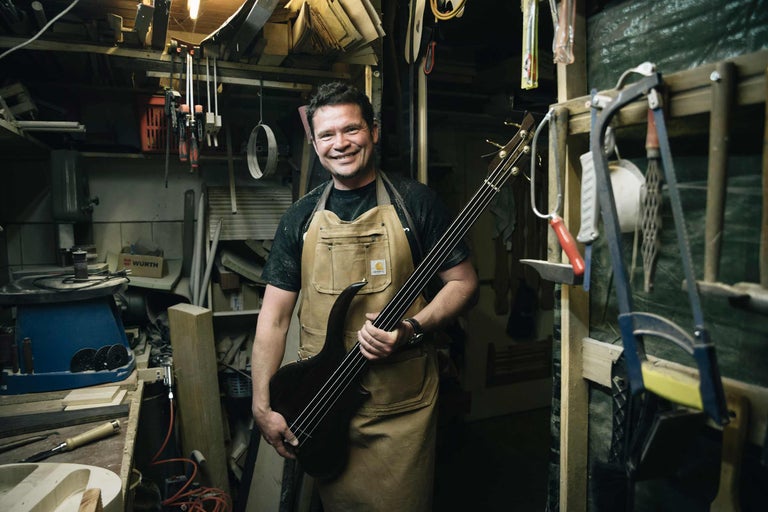 Ein Mann mit Carhartt-Schürze hält einen handgefertigten Bass in einer Werkstatt.
