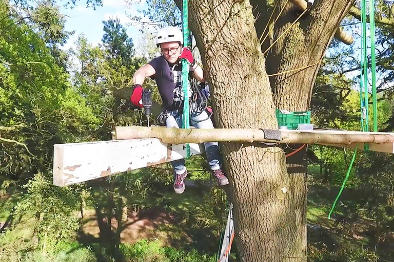 Mann mit Helm und Sicherheitsausrüstung arbeitet mit Bohrmaschine auf Baumplattform