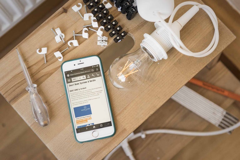 Szene mit Lampe, Lüsterklemmen, Schraubendreher, Kabelklemmen und Smartphone mit der Hornbach Webseite zum Thema Smart Home.