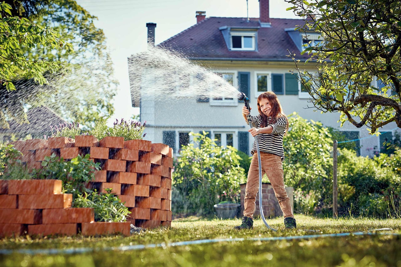 Mädchen bewässert Pflanzen in einem Garten mit einem Gartenschlauch und einer Gartenspritze.