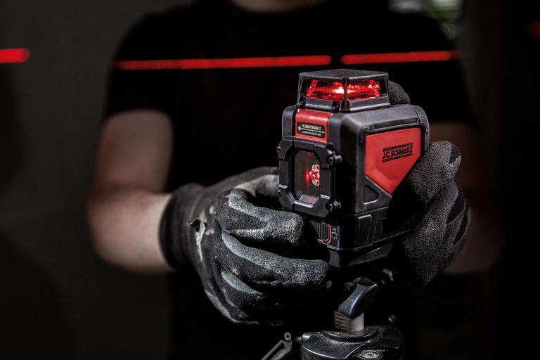 Person mit Arbeitshandschuhen hält einen J.C. Schwarz Kreuzlinienlaser auf einem Stativ, der rote Laserlinien projiziert.