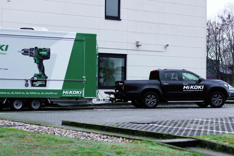 Ein schwarzer Pritschenwagen zieht einen Anhänger mit Werbung für Elektrowerkzeuge der Marke Hikoki.