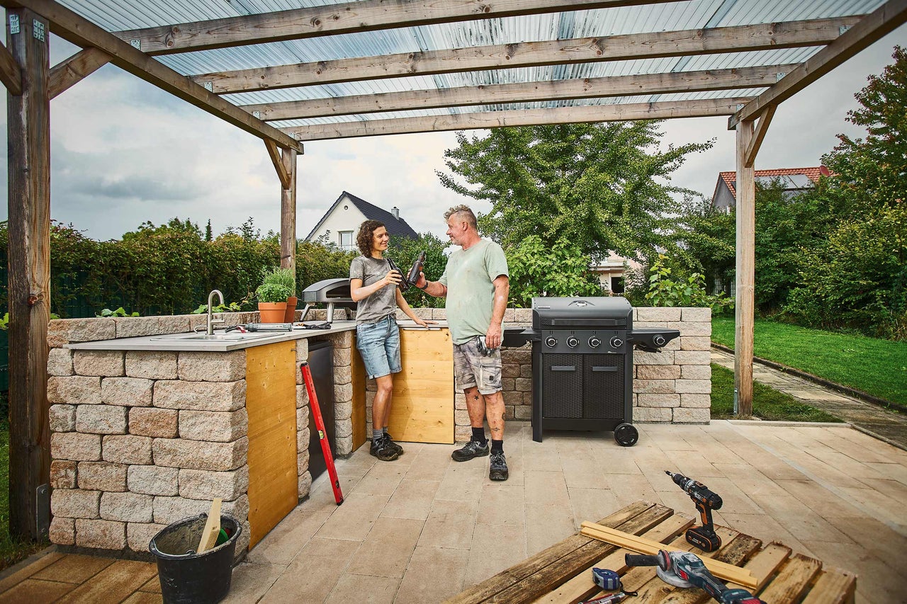 Außenküche aus Mauersteinen mit Gasgrill, Pizzaofen und Spülbecken unter einer Holzpergola, zwei Personen und Werkzeuge auf der Terrasse.