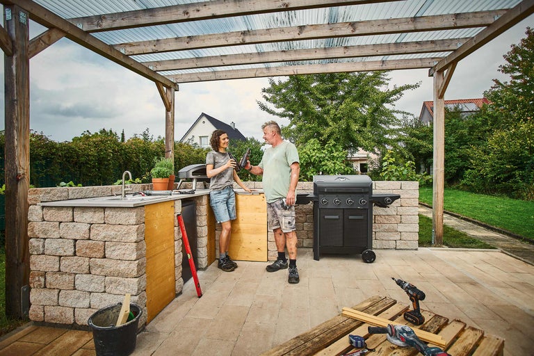 Außenküche aus Mauersteinen mit Gasgrill, Pizzaofen und Spülbecken unter einer Holzpergola, zwei Personen und Werkzeuge auf der Terrasse.