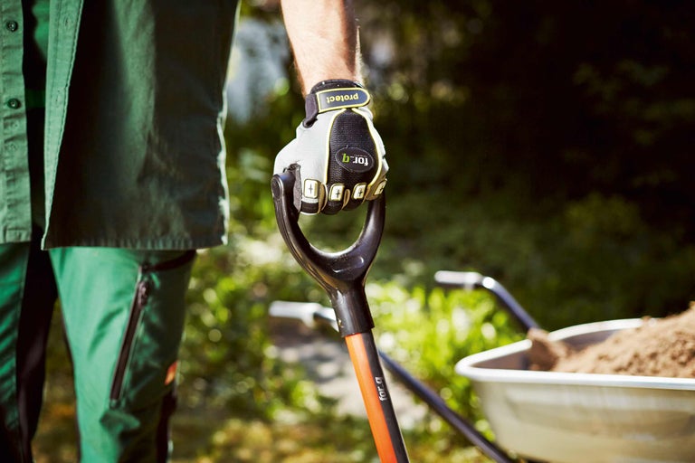 Eine Person mit Arbeitshandschuhen hält einen Spaten mit einem gefüllten Schubkarren im Hintergrund.