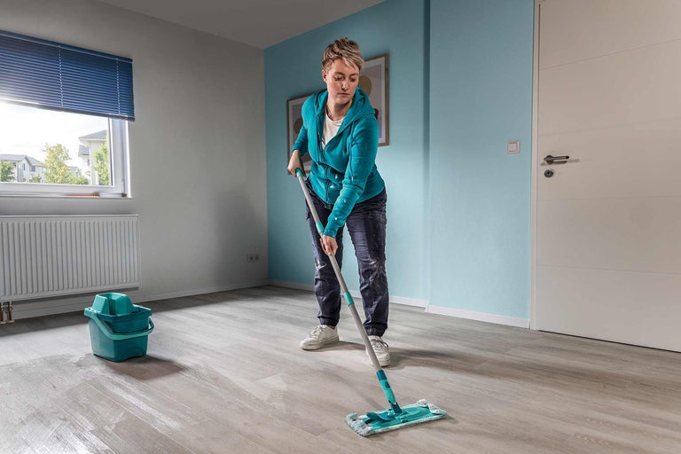 Frau wischt Boden mit Mopp in einem Zimmer mit Eimer und Heizkörper