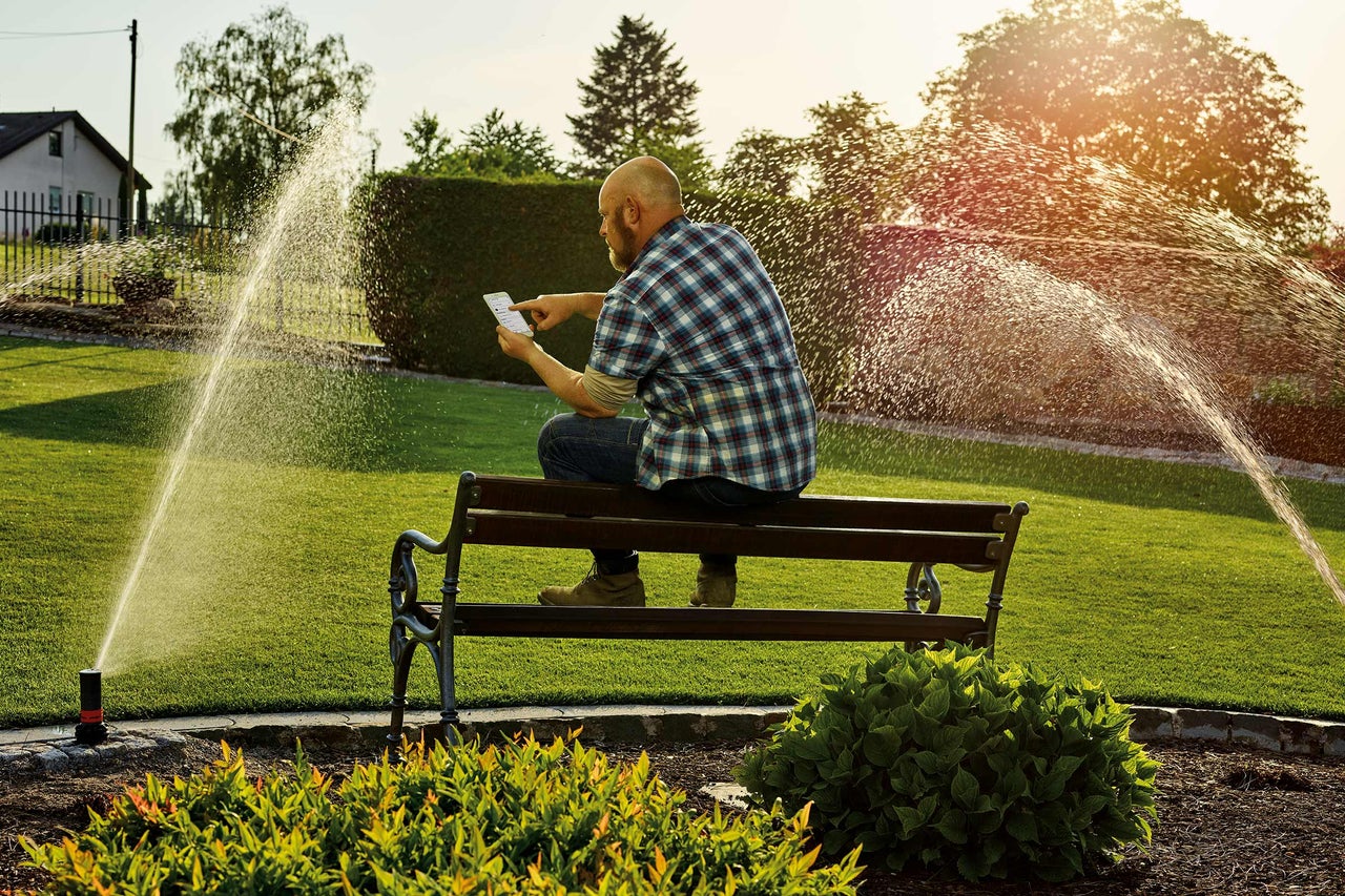 Mann sitzt auf einer Bank und bedient ein Bewässerungssystem per Smartphone in einem Garten.