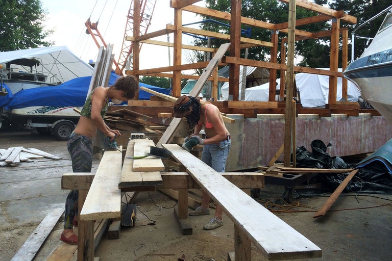 Zwei Personen arbeiten mit Elektrowerkzeugen an Holzkonstruktionen im Freien, im Hintergrund Boote und Gerüste
