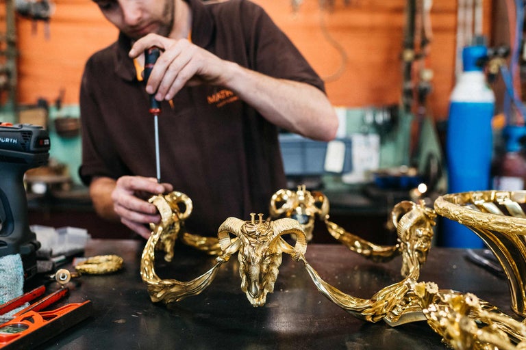 Ein Handwerker montiert ein Dekorationselement mit einem Schraubendreher. Das Element zeigt einen Widderkopf und ist aus Metall gefertigt.