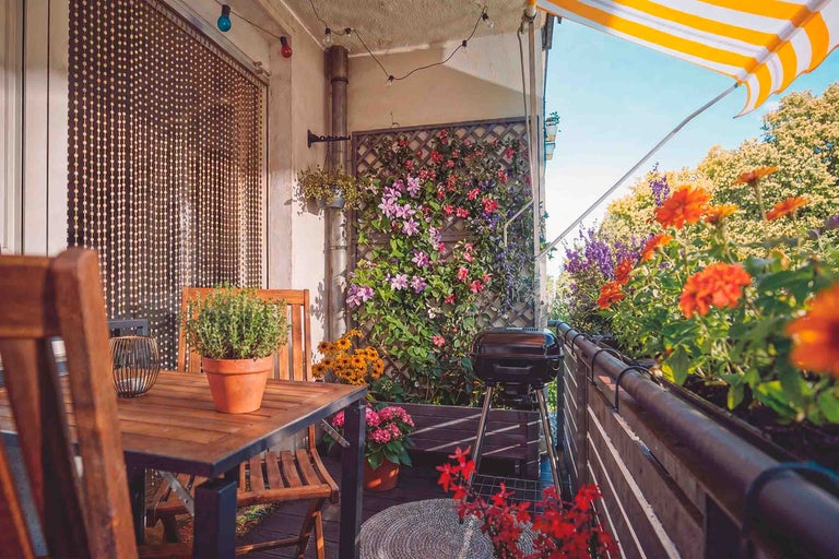 Gemütlicher Balkon mit Holzmöbeln, Kugelgrill, Blumen in Töpfen, Rankgitter mit Kletterpflanzen und gelb-weiß gestreifter Markise.