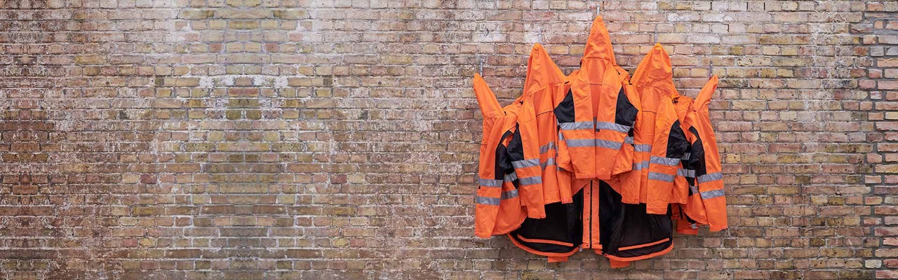 Mehrere Warnjacken mit Kapuze und Reflexstreifen hängen vor einer Backsteinmauer.
