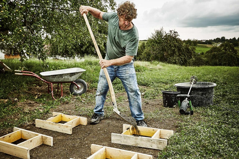 Ein Mann befüllt Holzrahmen mit einer Schaufel im Garten, während eine Schubkarre und ein Mörtelkübel bereitstehen.