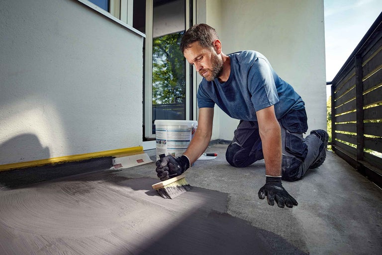 Mann trägt mit einem Pinsel eine graue Beschichtung auf einen Balkonboden auf