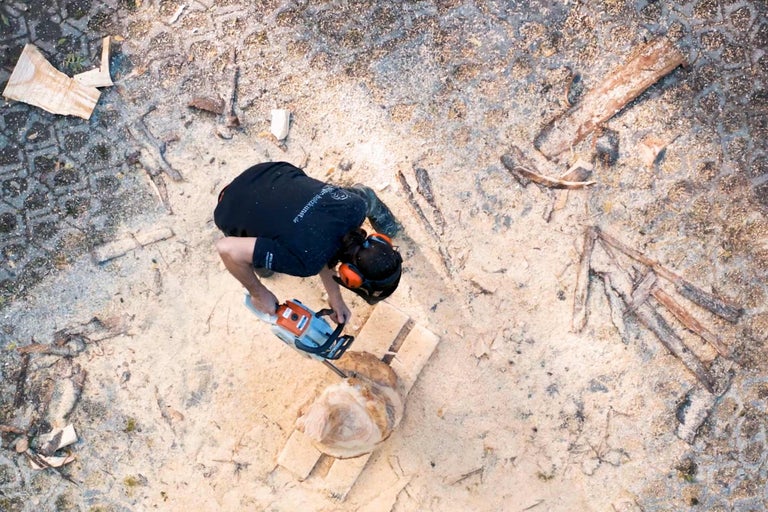 Draufsicht einer Person, die mit einer Kettensäge ein Stück Holz bearbeitet, umgeben von Sägemehl.