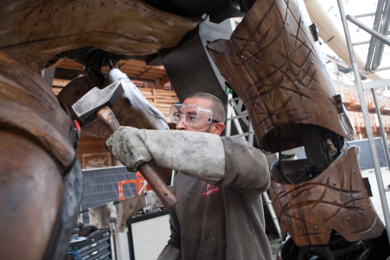 Ein Handwerker mit Schutzbrille bearbeitet mit einem Hammer ein großes Holzobjekt.