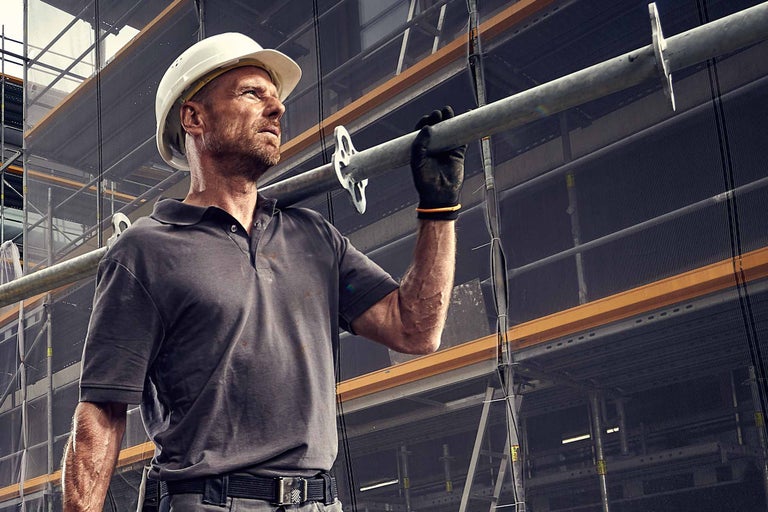 Bauarbeiter mit Schutzhelm trägt Gerüststange vor Baugerüst