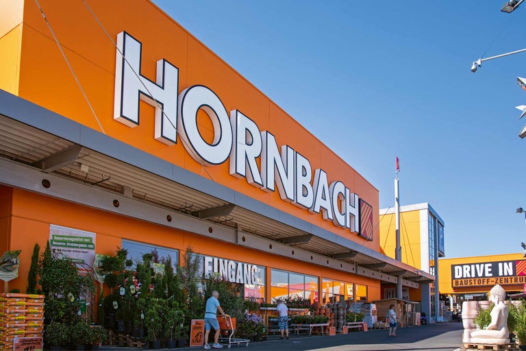 Außenansicht eines Hornbach Baumarktes mit Pflanzen und Einkaufswagen vor dem Eingang.