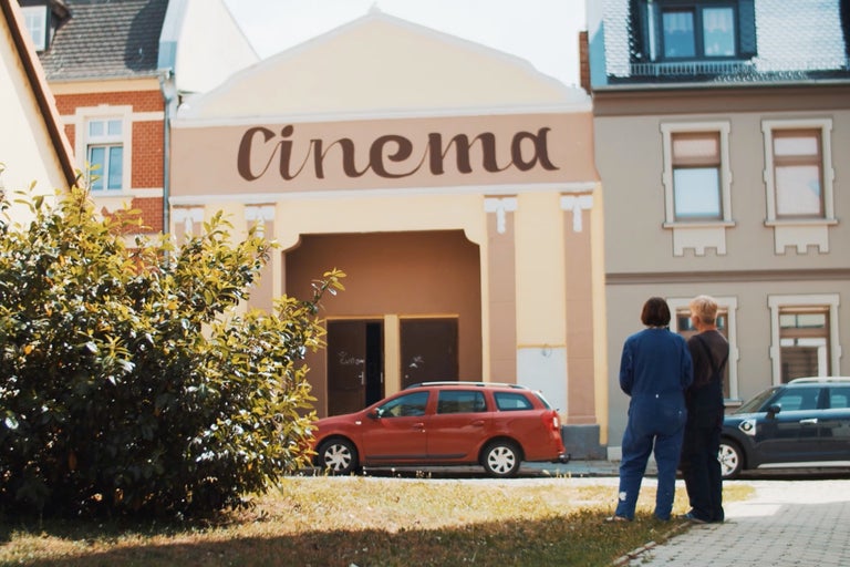 Außenansicht eines Kinos mit zwei Personen und Autos vor dem Gebäude