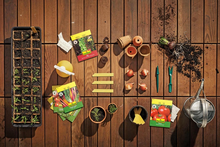 Gartenszene mit Anzuchtschale, Saatguttüten von FloraSelf, Pflanztöpfen und Gartenwerkzeug auf Holzboden.