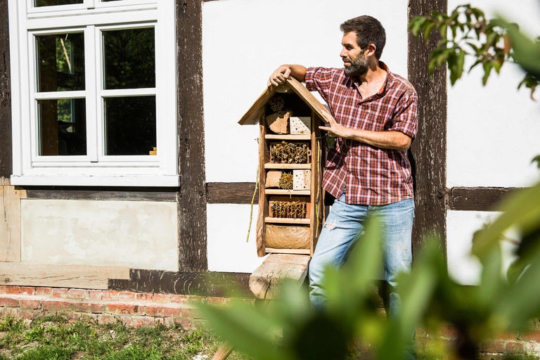 Mann lehnt an einem Insektenhotel im Garten vor einem Fachwerkhaus.