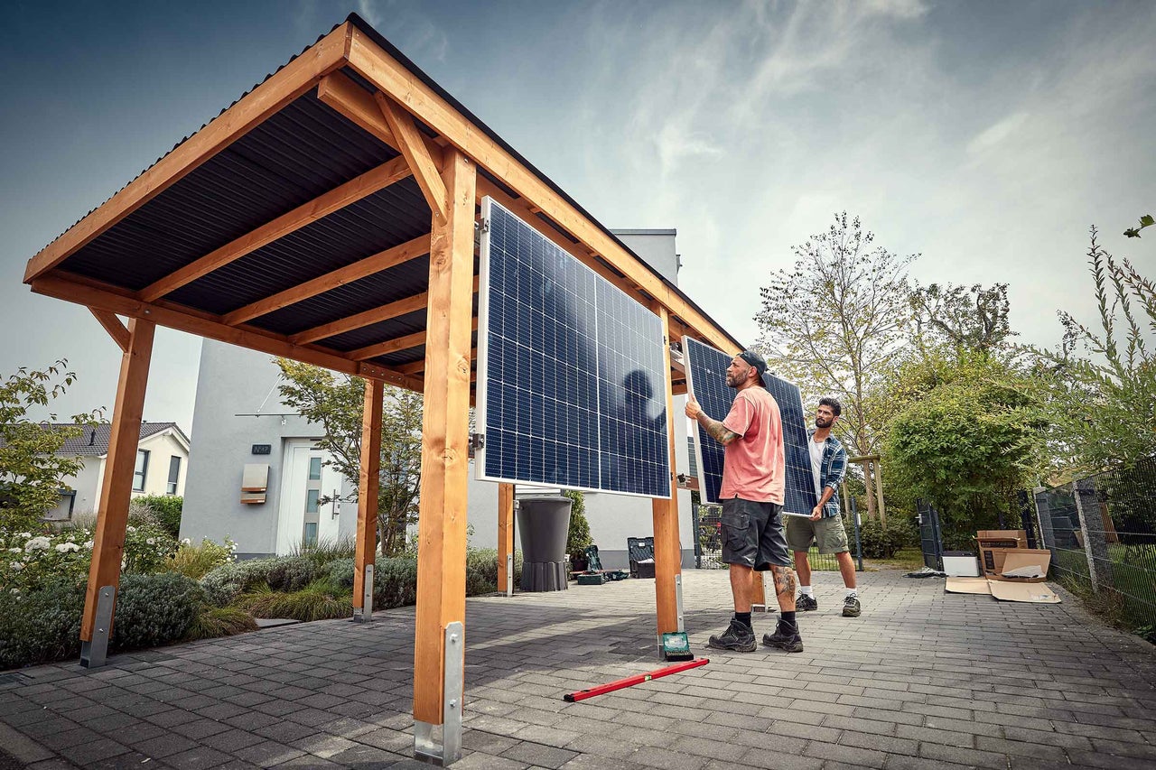 Zwei Personen montieren Solarmodule an einem hölzernen Carport mit dunklem Wellblechdach auf einer gepflasterten Fläche vor einem Wohnhaus.