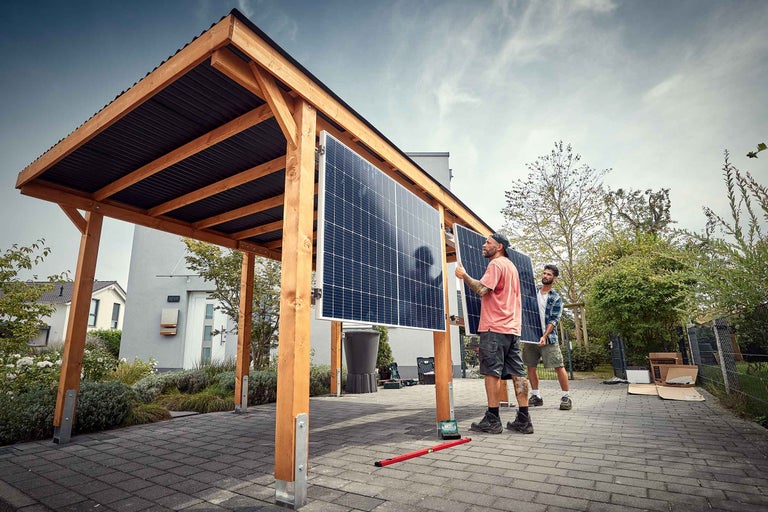 Zwei Personen montieren Solarmodule an einem hölzernen Carport mit dunklem Wellblechdach auf einer gepflasterten Fläche vor einem Wohnhaus.