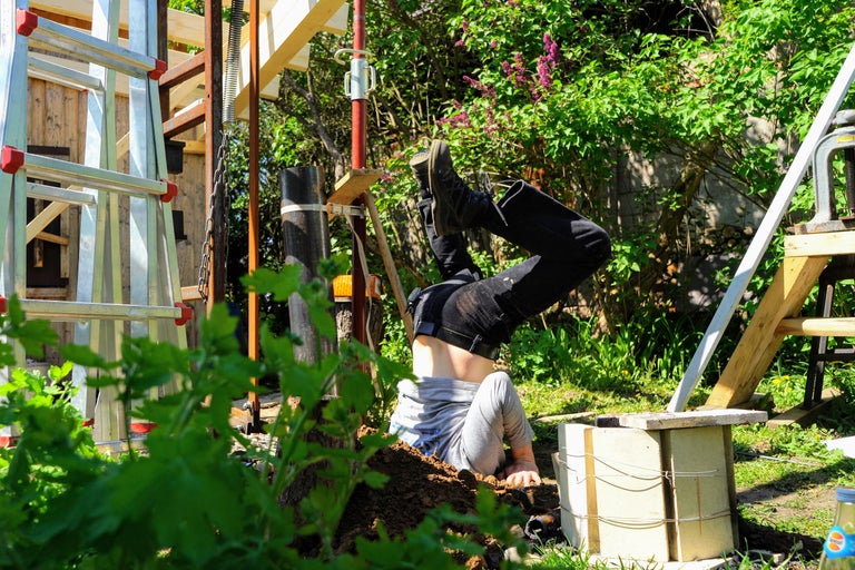 Eine Person, die kopfüber in einem Loch steckt, umgeben von Bauwerkzeugen und Pflanzen.