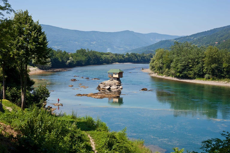 Haus auf einem Felsen im Fluss mit Person, umgeben von Bäumen und Bergen