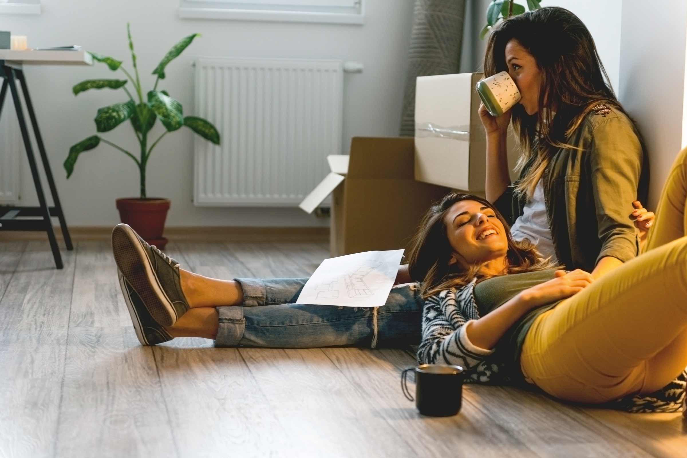 Zwei Frauen entspannen sich in einem hellen Raum mit Laminatboden, Umzugskartons, Zimmerpflanze, Heizkörper, Tisch und Tasse Kaffee