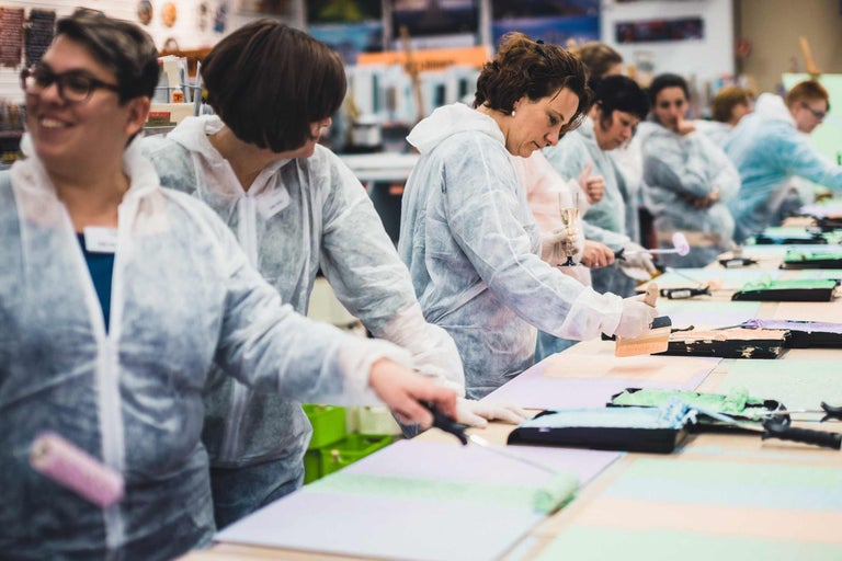 Mehrere Personen arbeiten mit Farbe, Pinseln und Farbrollern an Tischen in einem hellen Raum.